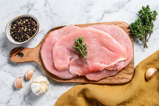 2 raw turkey breast steak stacked on top of each other on a wooden board with a kitchen towel and herbs and spices 